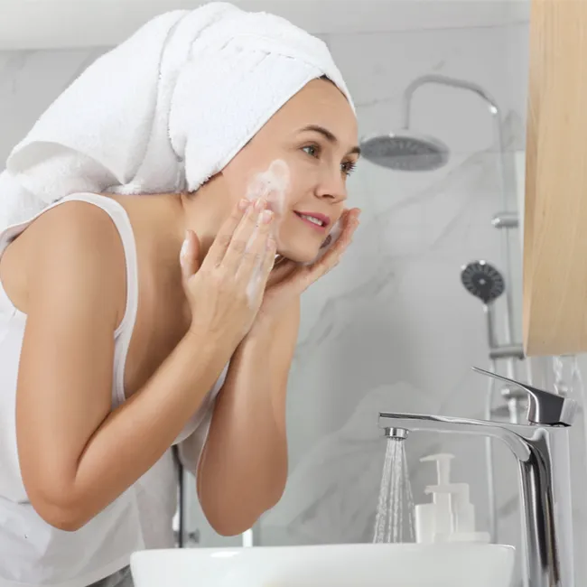 woman white towel hair wrap washing face with soap and water over sink looking in mirror